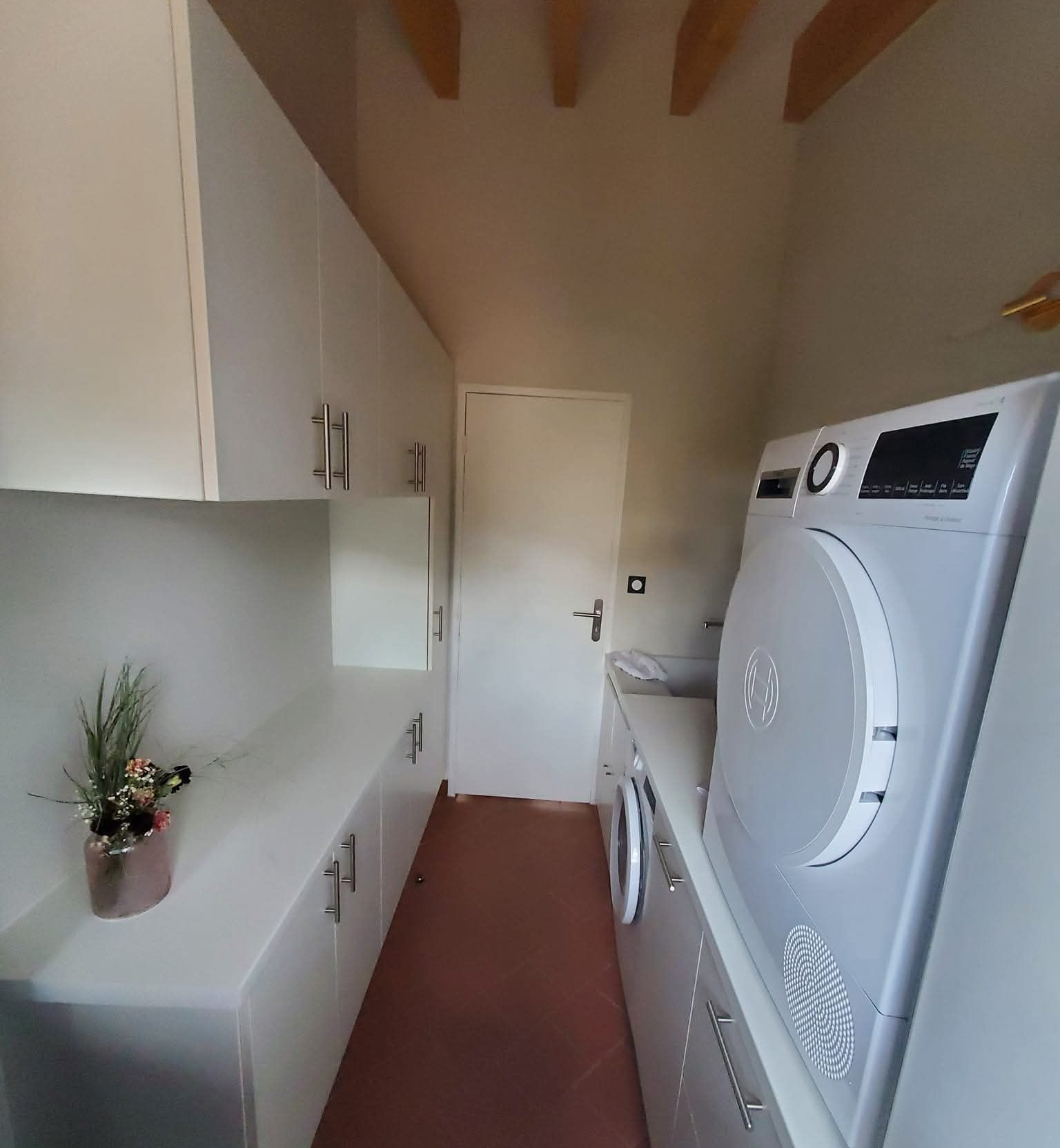 Buanderie avec placards blancs, sèche-linge et lave-linge. Une porte se trouve au fond de l'espace étroit.
