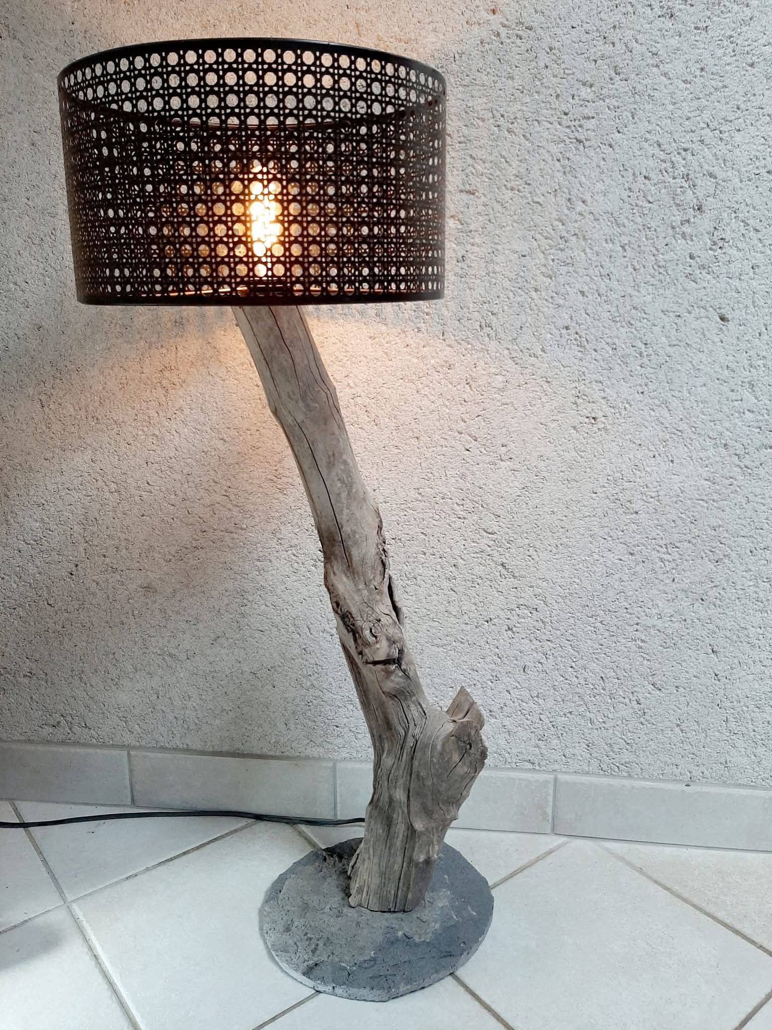 Lampadaire avec un abat-jour noir tissé et une base de style en bois flotté sur une base circulaire grise.