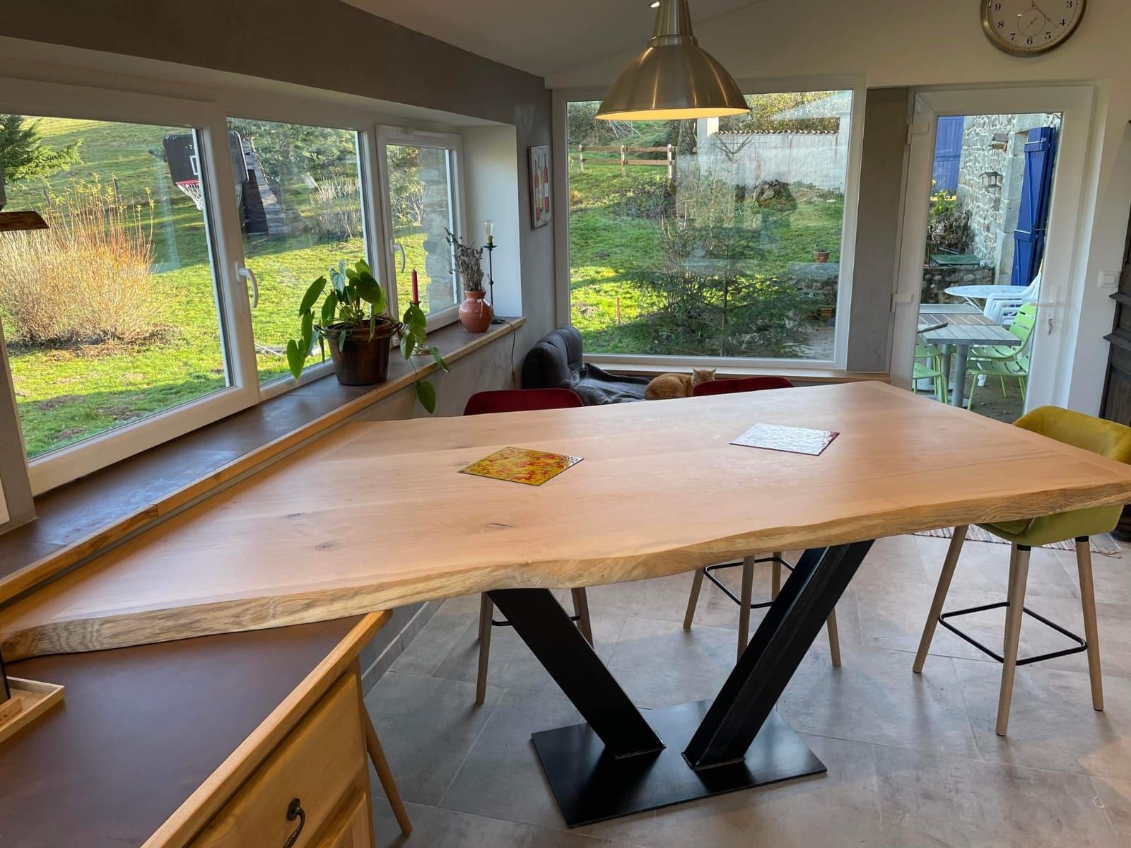Table en bois clair avec base en métal noir dans une salle à manger lumineuse, avec des fenêtres donnant sur une cour.