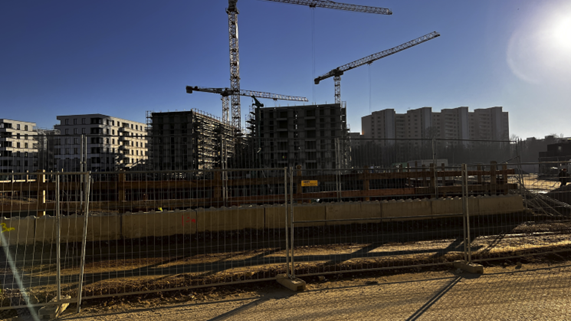 Eine Fotografie einer Großbaustelle mit Bauzäunen im Vordergrund und Kränen vor Rohbauten im Hintergrund.