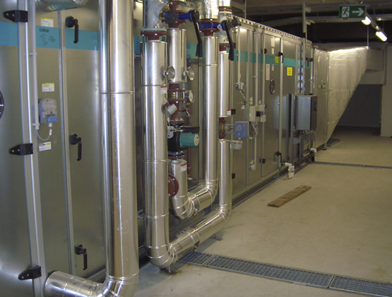 Eine Fotografie von metallischen Industrie-Lüftungsgeräten und isolierten Rohrleitungen in einem Technikraum.