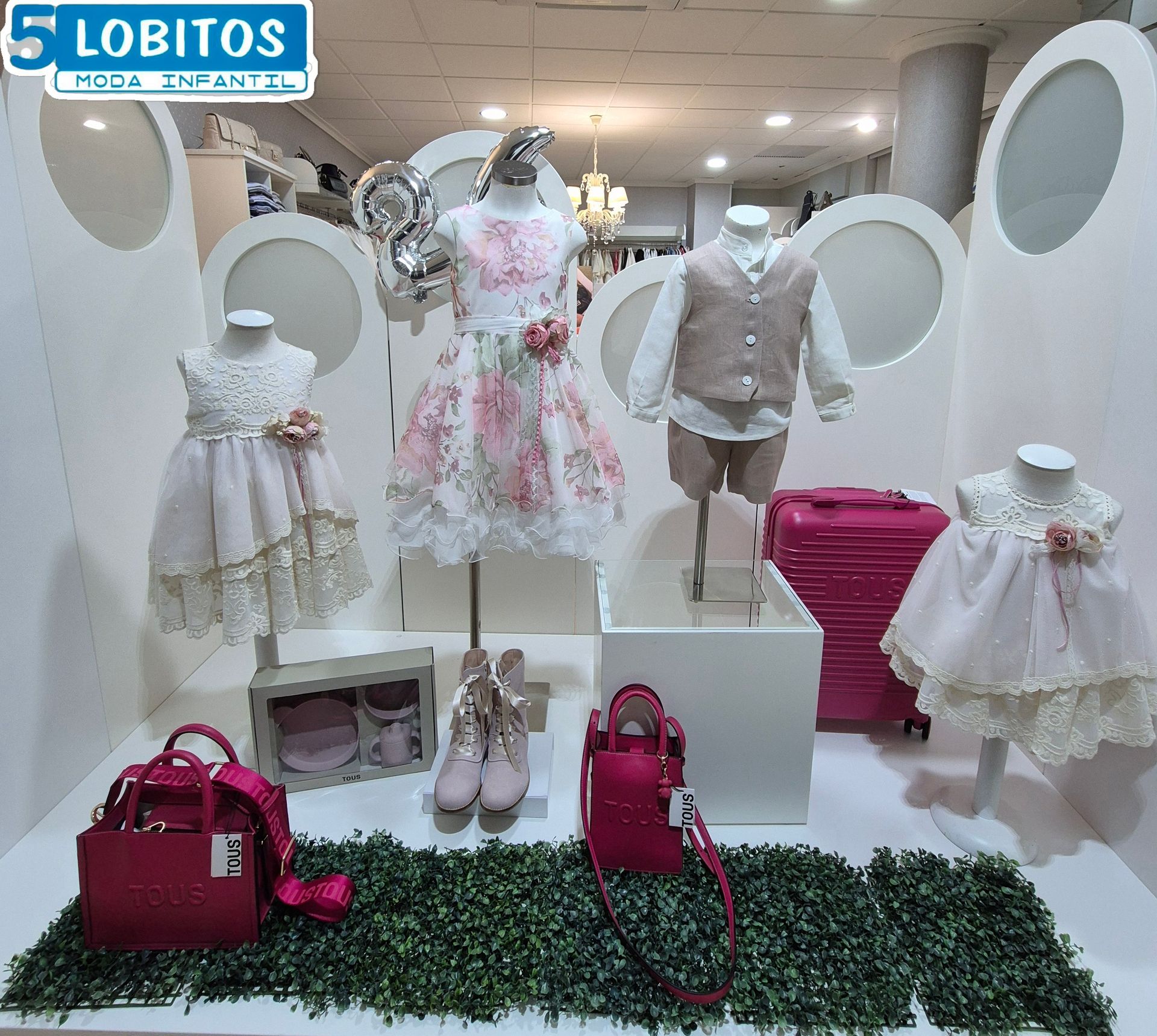 Un escaparate con ropa formal infantil. Vestidos blancos y rosas, trajes beige y accesorios rosas se presentan sobre maniquíes.