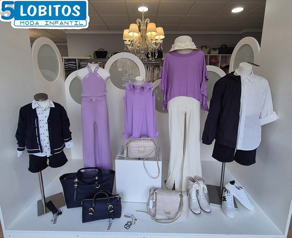 Un escaparate con ropa infantil en tonos morados, blancos y azul marino. Varios maniquíes muestran conjuntos con bolsos y zapatos.