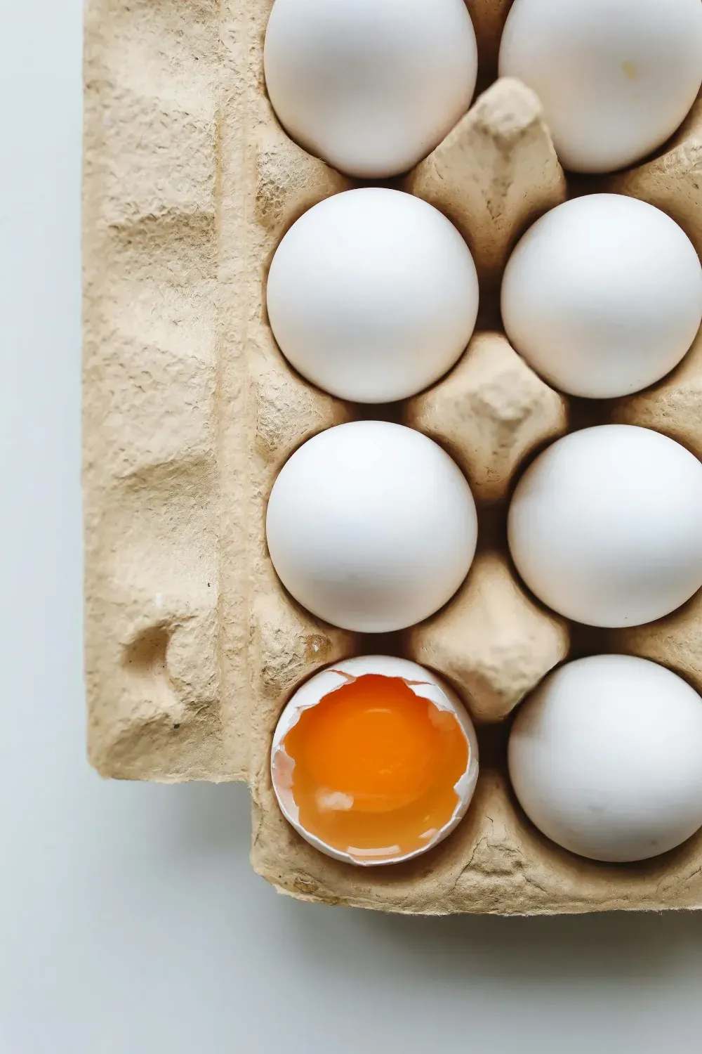 Caja de huevos blancos con un huevo roto que deja ver la yema de color naranja brillante.
