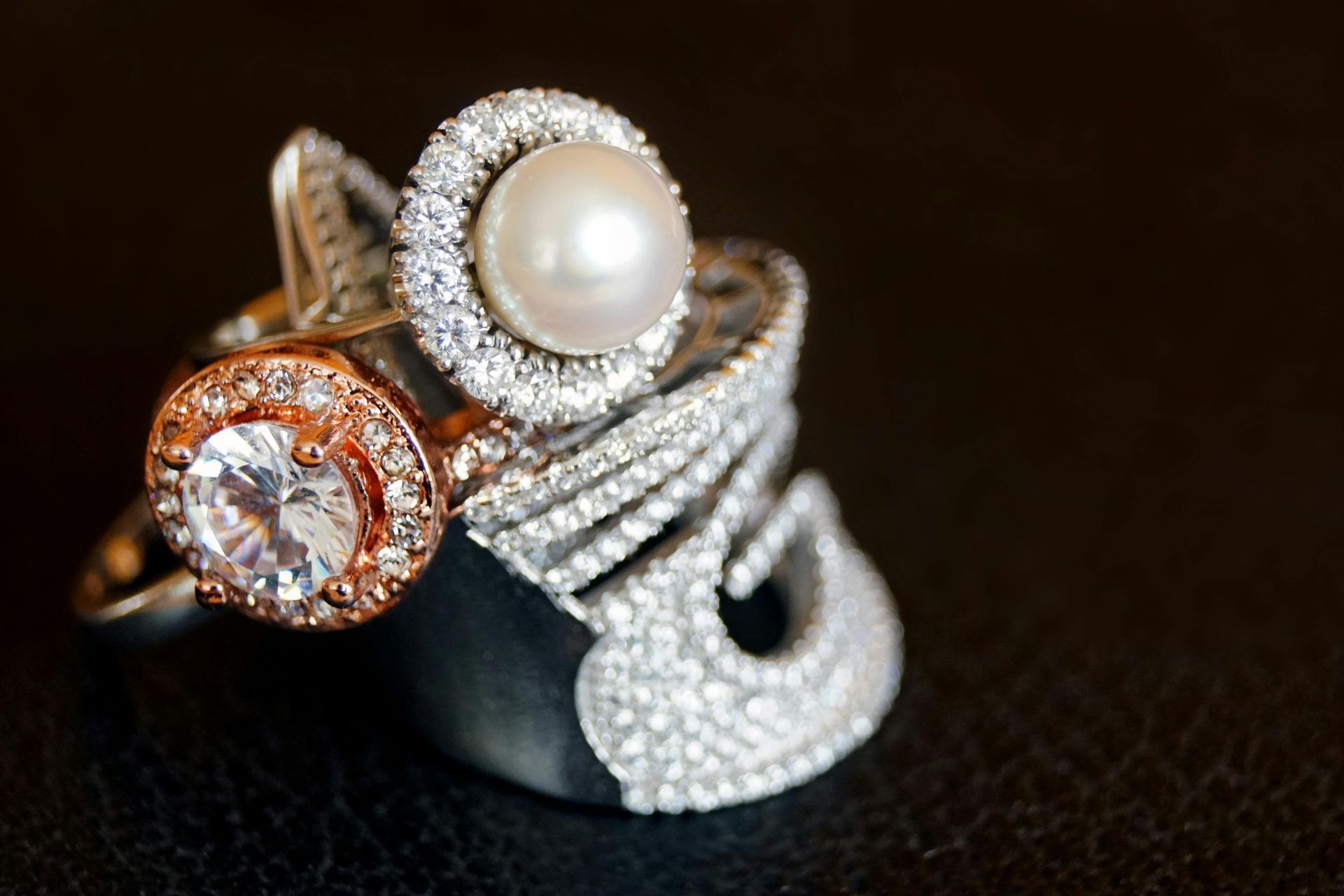 Tres anillos enjoyados con perlas, diamantes y detalles de oro rosa sobre una superficie oscura.