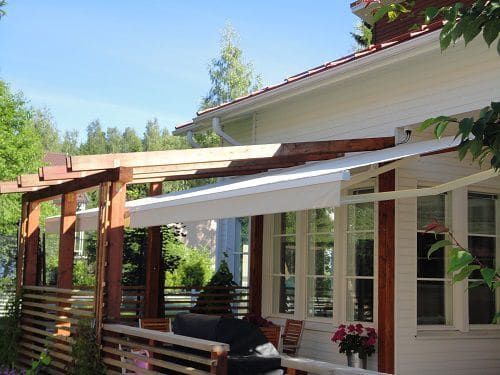 Puinen pergola kiinnitettynä beigenväriseen taloon, jossa on valkoinen markiisi ja roikkuvat kukkakorit sinisen taivaan alla