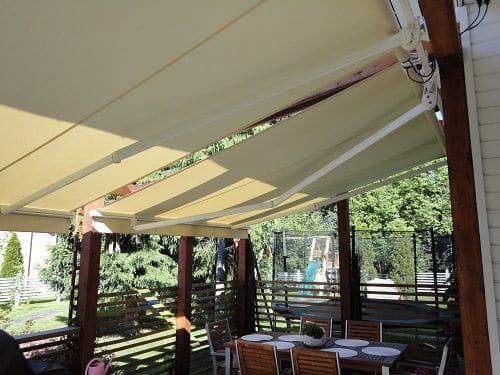 Katettu patio beigenvärisillä markiiseilla, puisilla pöydillä ja ulkona puutarhan vehreydellä
