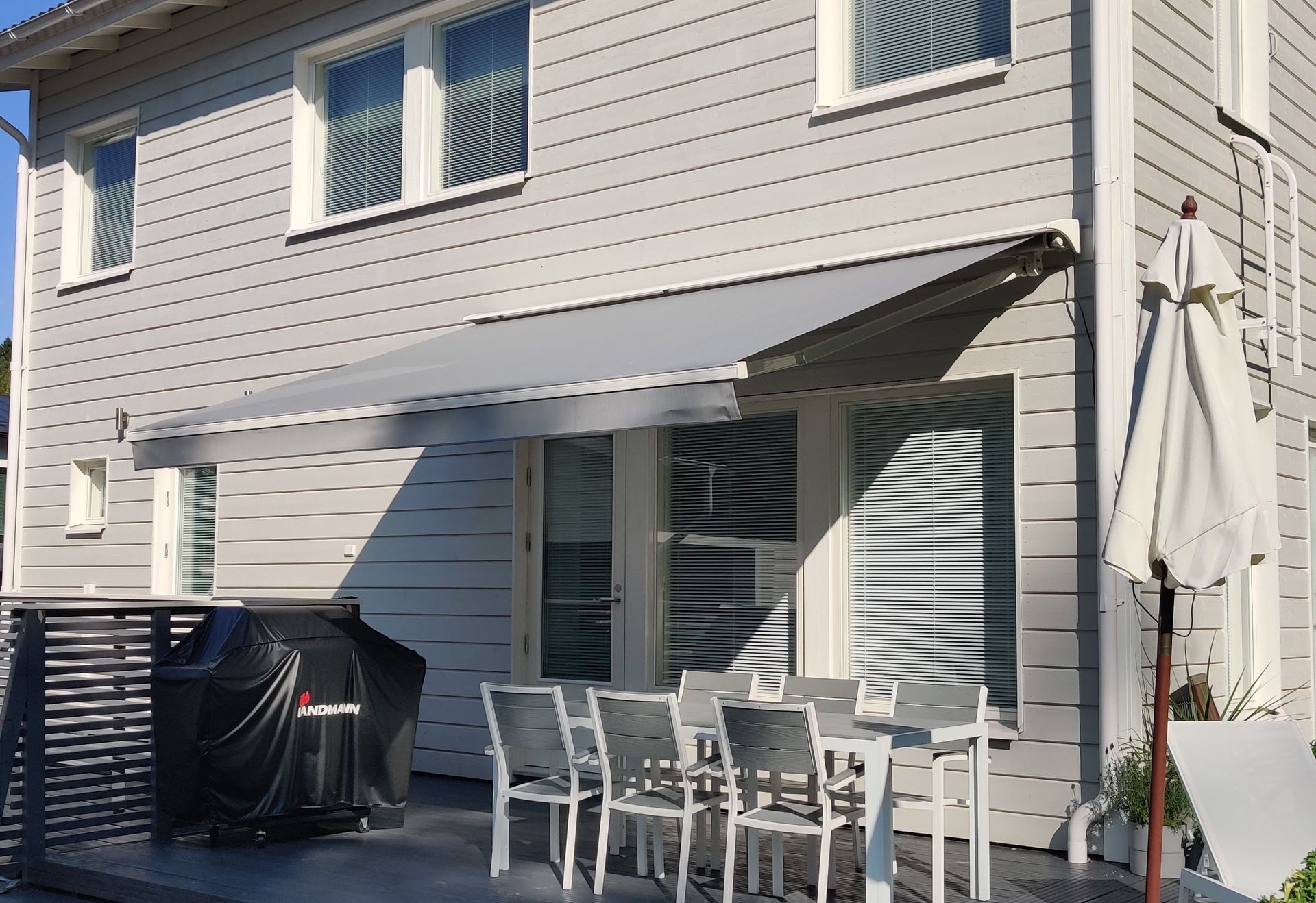Patio, jossa on valkoinen ulkoruokailuryhmä, grilli ja suljettu aurinkovarjo beigenvärisen talon vieressä, jossa on raidallinen markiisi.