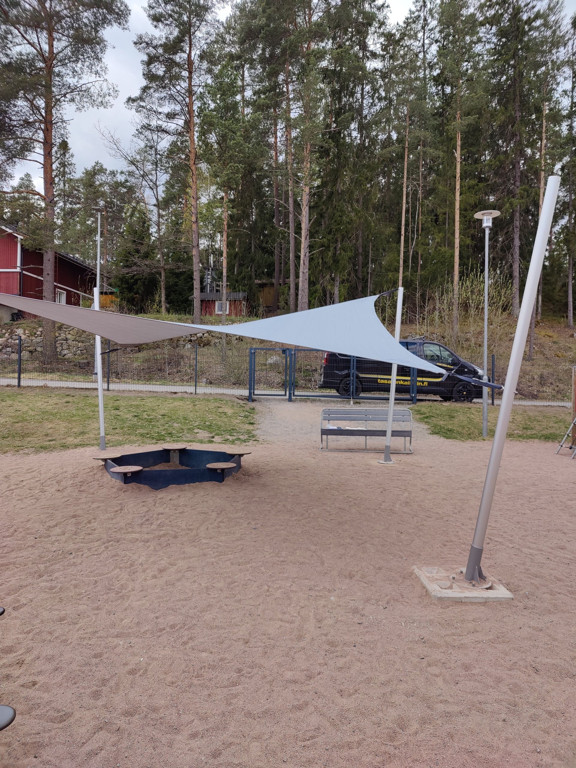 Ulkoleikkikenttä, jossa on varjopurjeita, sorakenttä ja penkkejä korkeiden puiden vieressä.