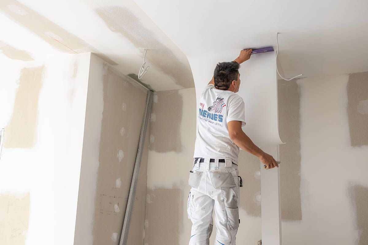 Ein Mann auf einer Leiter trägt in einem im Bau befindlichen Raum Spachtelmasse auf Trockenbauwände auf.