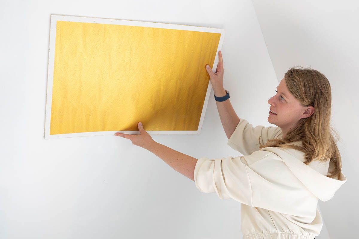 Frau hält ein goldfarbenes Kunstwerk und ist dabei, es an eine weiße Wand in einem Raum zu hängen.