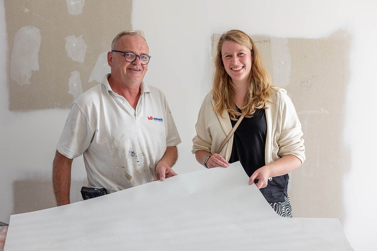 Ein Mann und eine Frau lächeln und halten ein weißes Laken vor einer Wand, die gerade renoviert wird.
