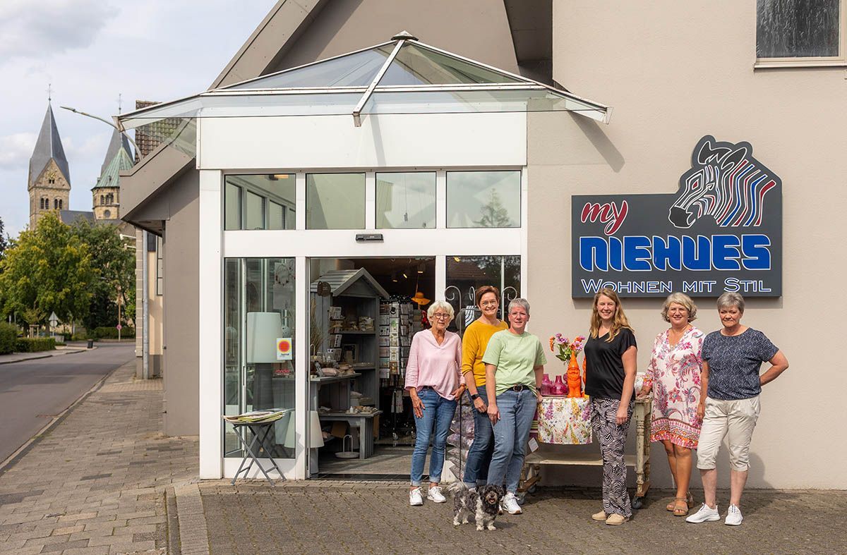 Sechs lächelnde Frauen vor einem Geschäft namens „Mehus“. Beiges Gebäude, Glastür, Kirche im Hintergrund.