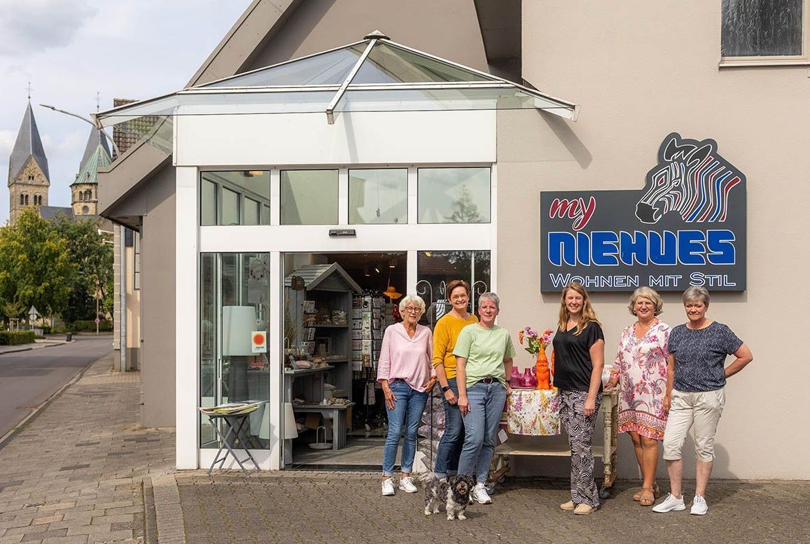 Gruppe von Frauen vor einem Geschäft namens „NEUES“ mit Glastüren, beigen Wänden und einem Kirchturm im Hintergrund.