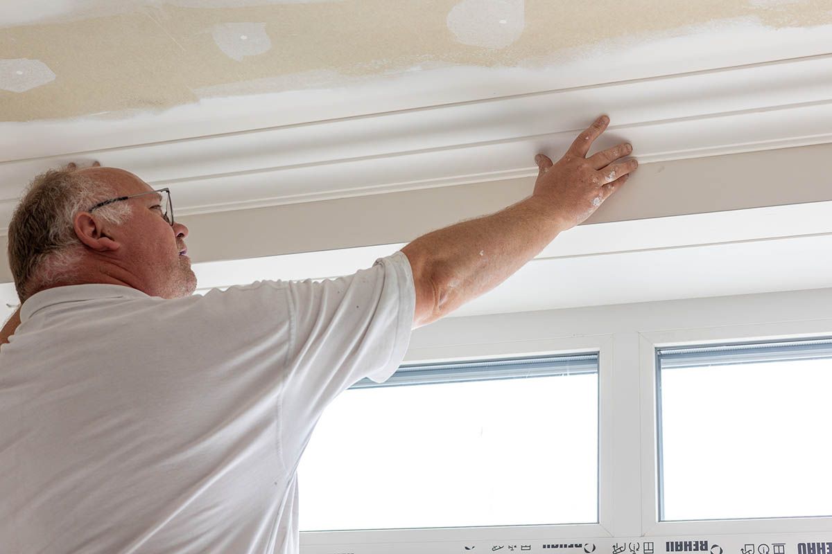 Mann installiert weiße Zierleisten an einer Decke in der Nähe eines Fensters.