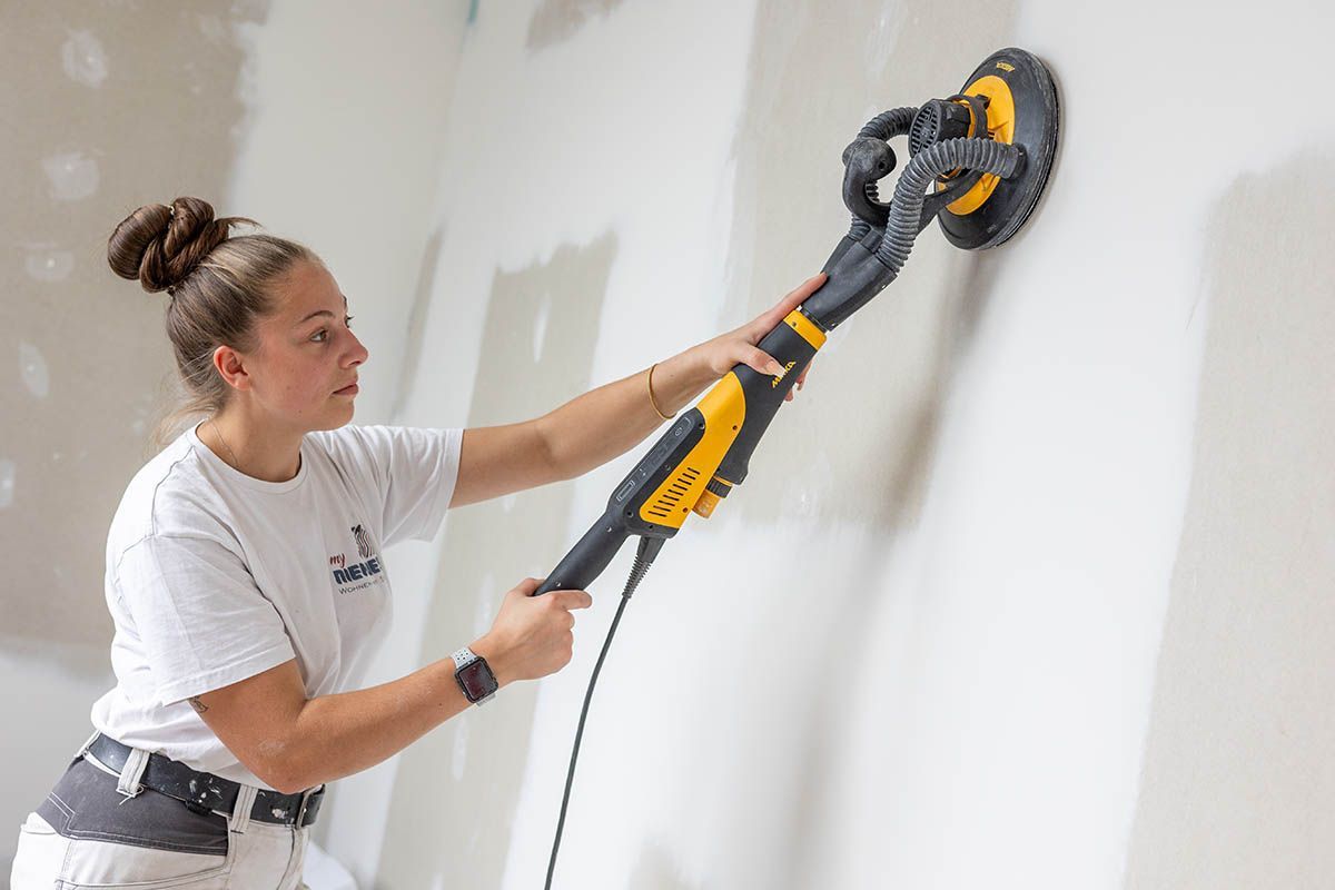 Frau schleift in einem Innenbereich eine Wand mit einem elektrischen Schleifgerät. Sie trägt ein T-Shirt und hat ihr Haar hochgesteckt.