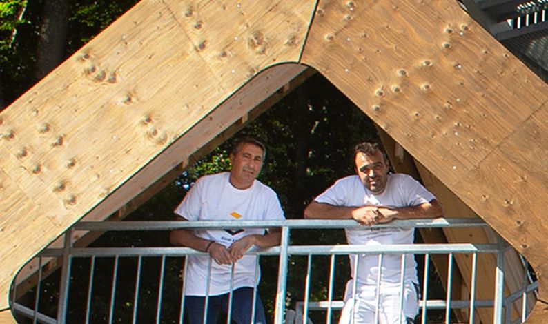 Zwei Männer stehen auf einem Balkon mit einer Holzkonstruktion im Hintergrund