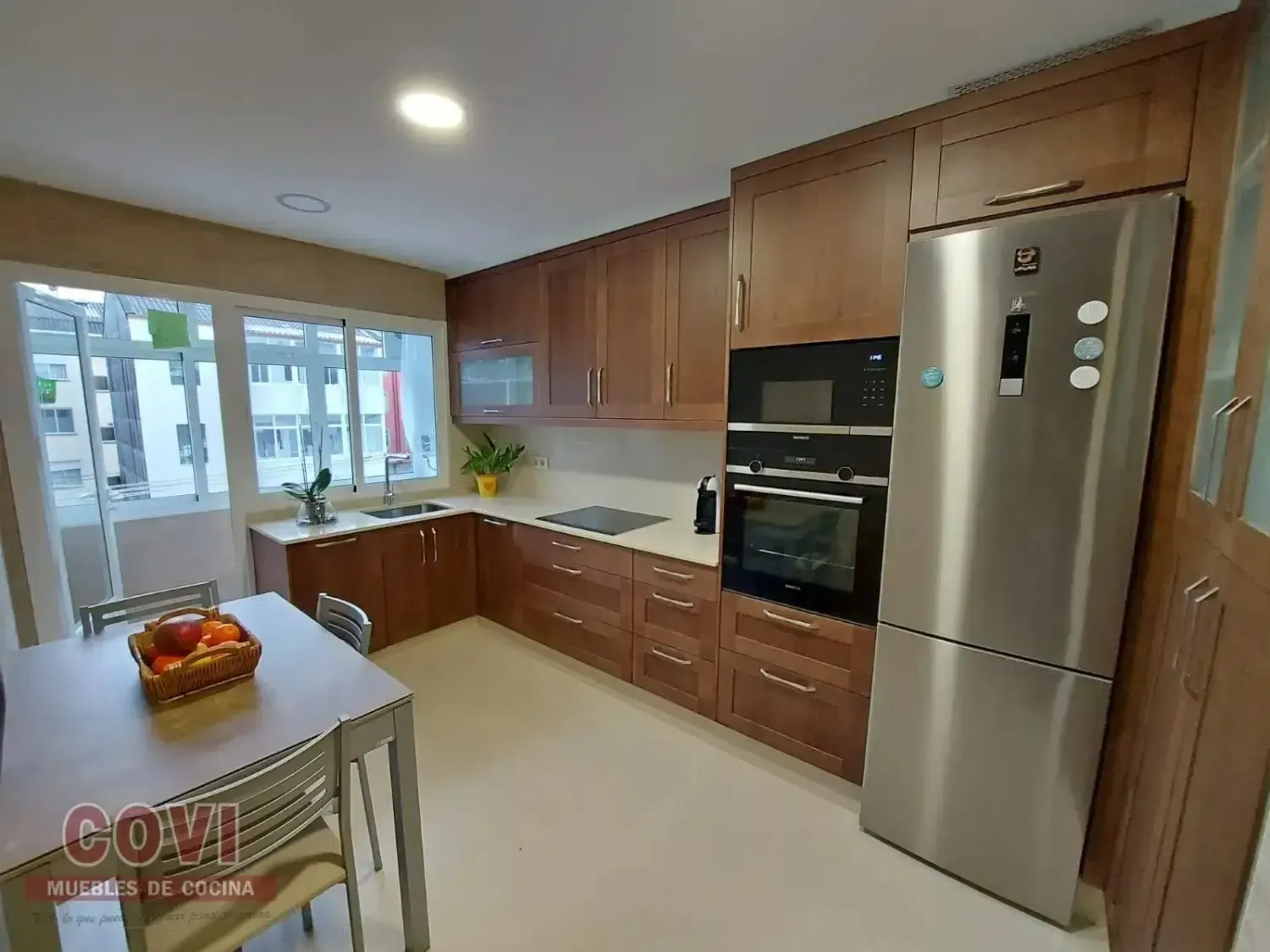 Cocina en forma de L con gabinetes de madera, refrigerador de acero inoxidable, encimeras blancas y pequeña mesa de comedor.