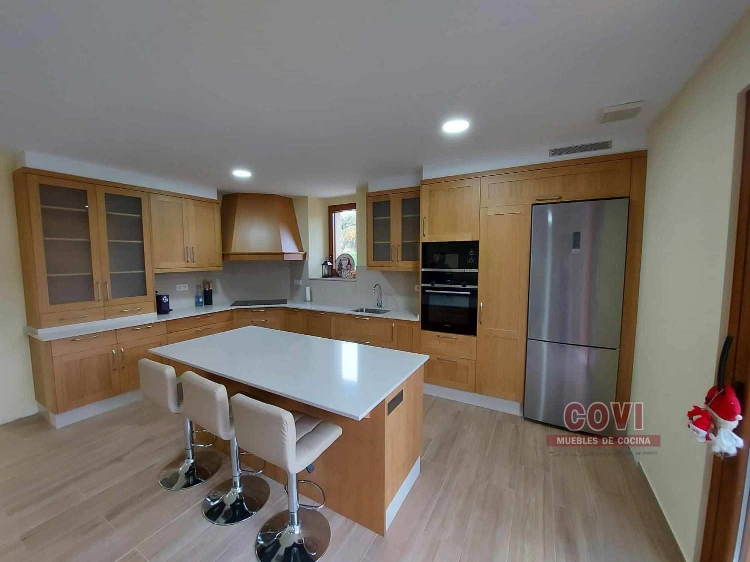 Cocina moderna con gabinetes de madera clara, refrigerador de acero inoxidable e isla con taburetes de bar.