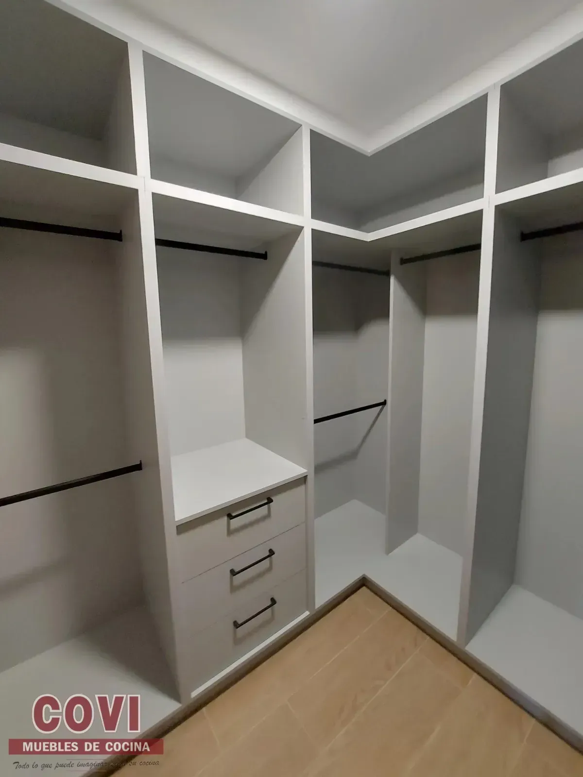 Armario empotrado blanco con estantes, cajones y barras para colgar. Esquinero, con suelo de madera a la vista.