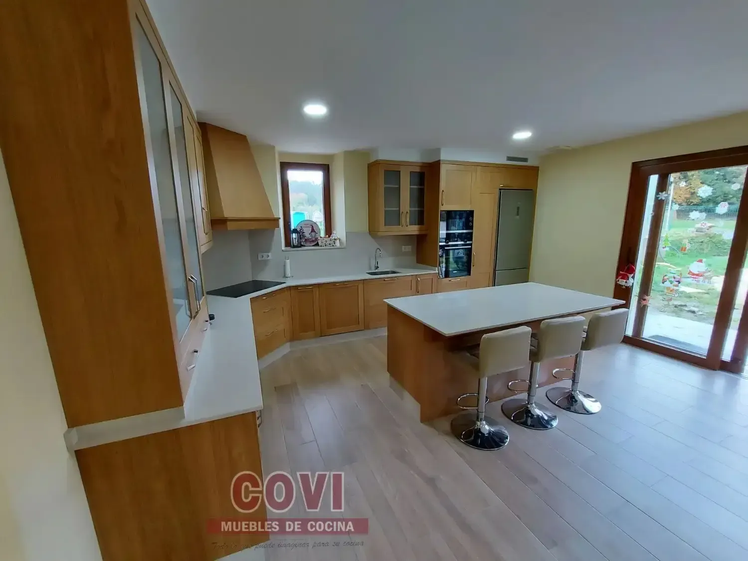 Una cocina de madera clara con gabinetes, una isla central con asientos y ventanas grandes.