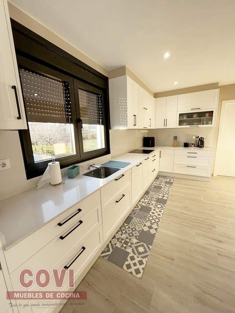 Cocina blanca moderna con encimeras largas, ventana negra y alfombra estampada.
