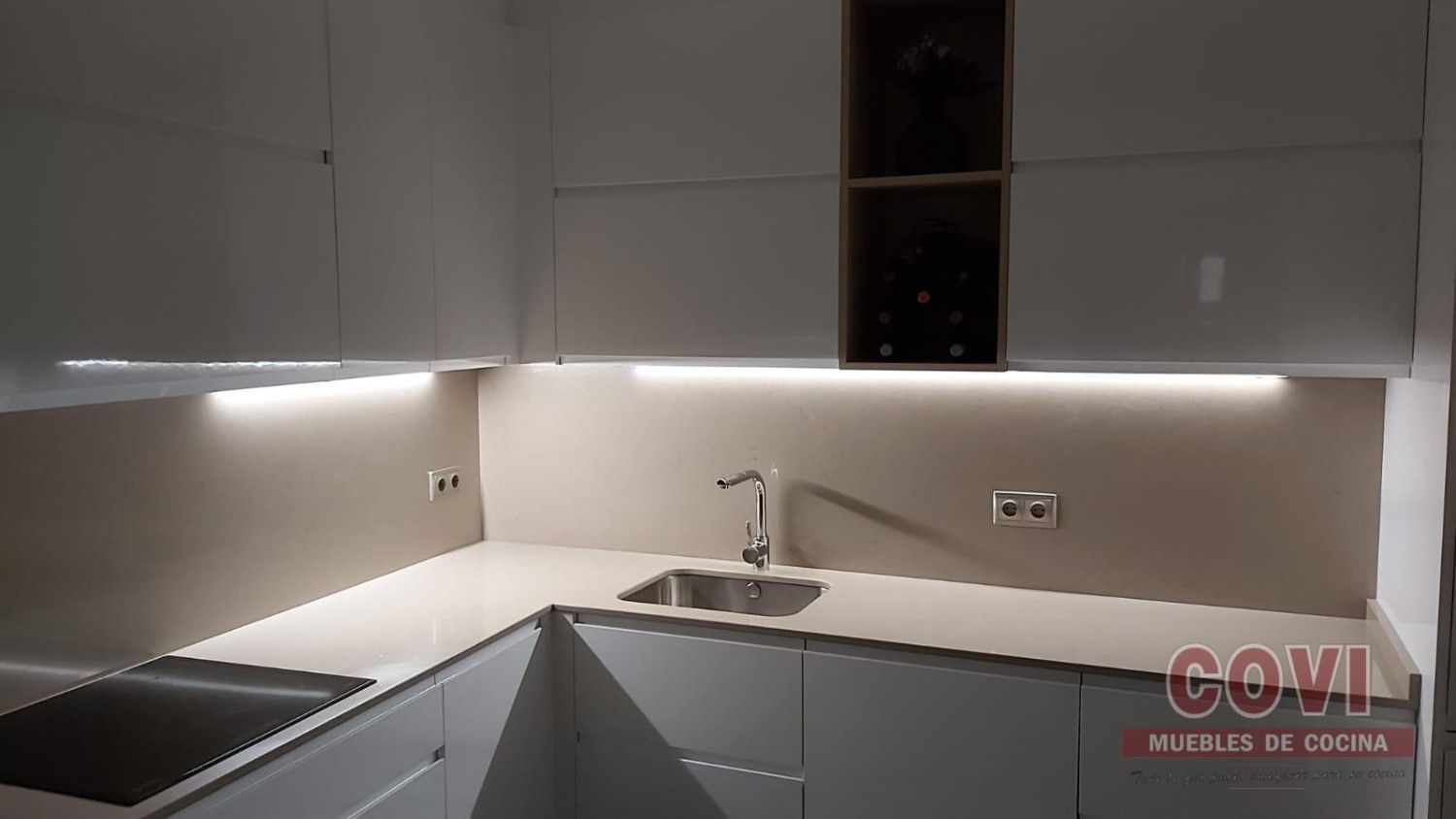 Cocina moderna y blanca con iluminación debajo de los gabinetes, fregadero de acero inoxidable y estufa incorporada.
