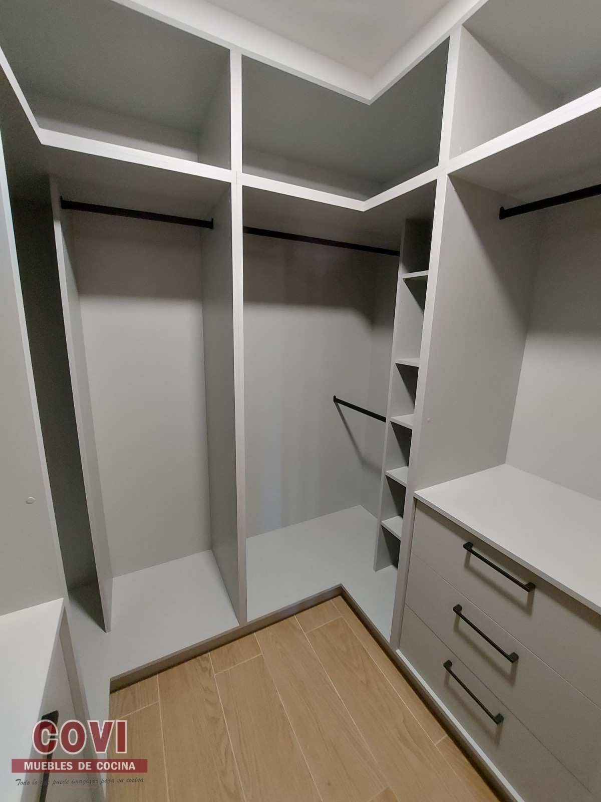 Vestidor con estantes, cajones y suelo de madera en gris claro. Barras y tiradores negros para colgar.
