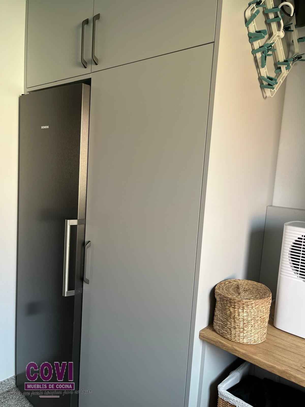 Armarios grises con un refrigerador negro. Se ven una cesta de mimbre y un pequeño estante.