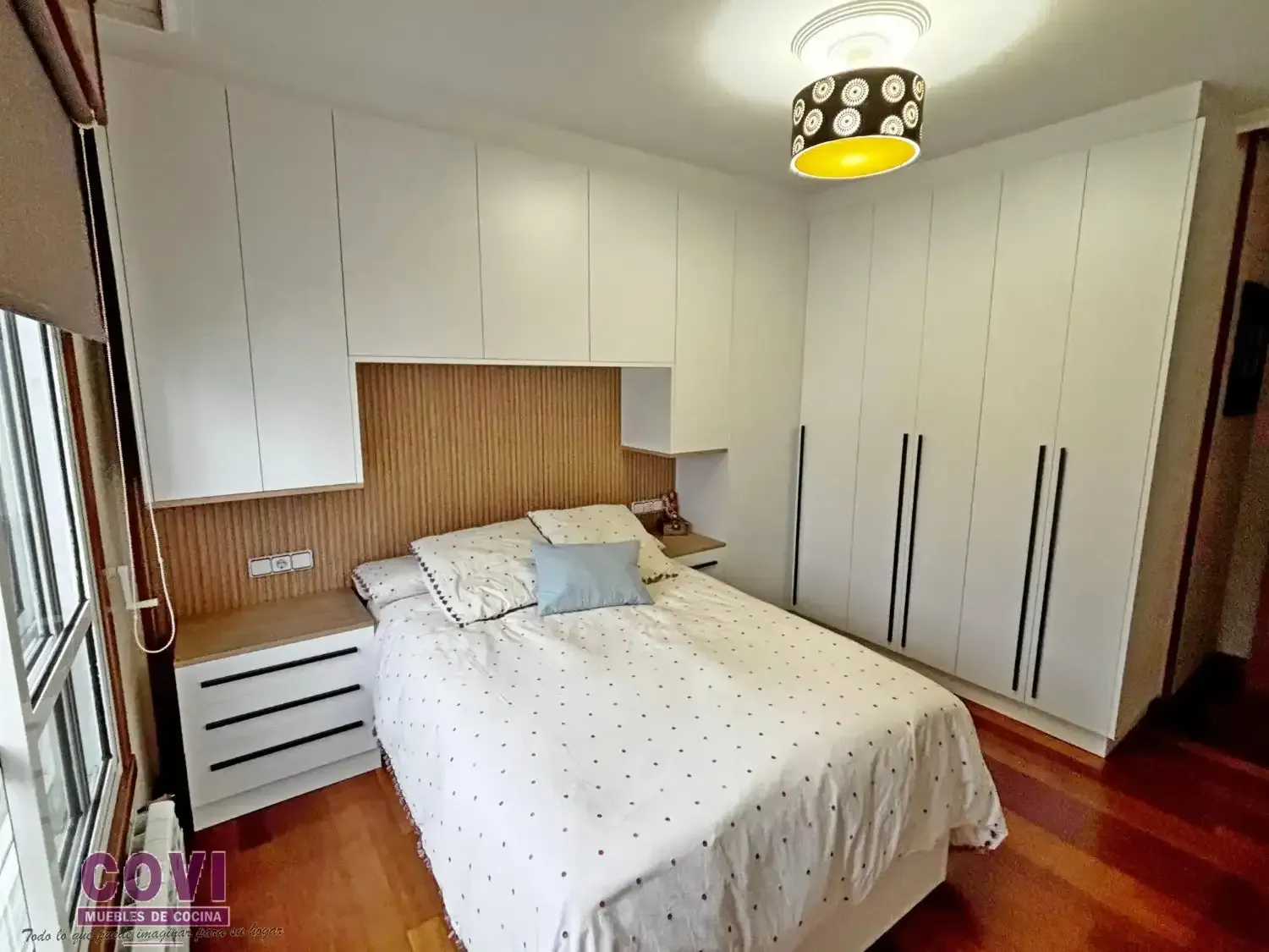 Dormitorio con muebles empotrados blancos, cama con almohada azul, cabecero de madera y suelo de madera.