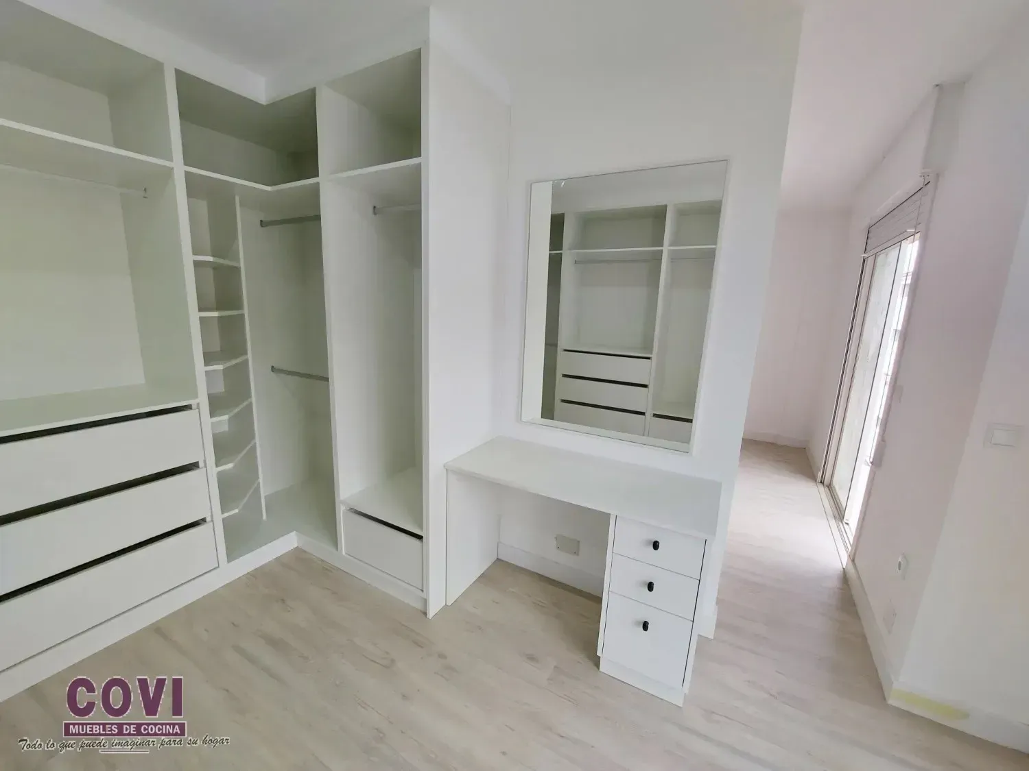 Vestidor blanco con tocador, espejo, cajones y barras para colgar. Suelo de madera clara.