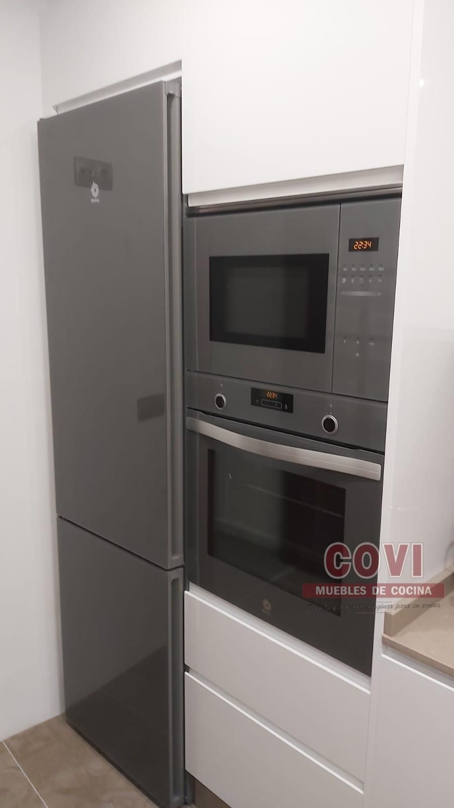 Electrodomésticos de cocina empotrados: refrigerador gris, microondas y horno dentro de gabinetes blancos.