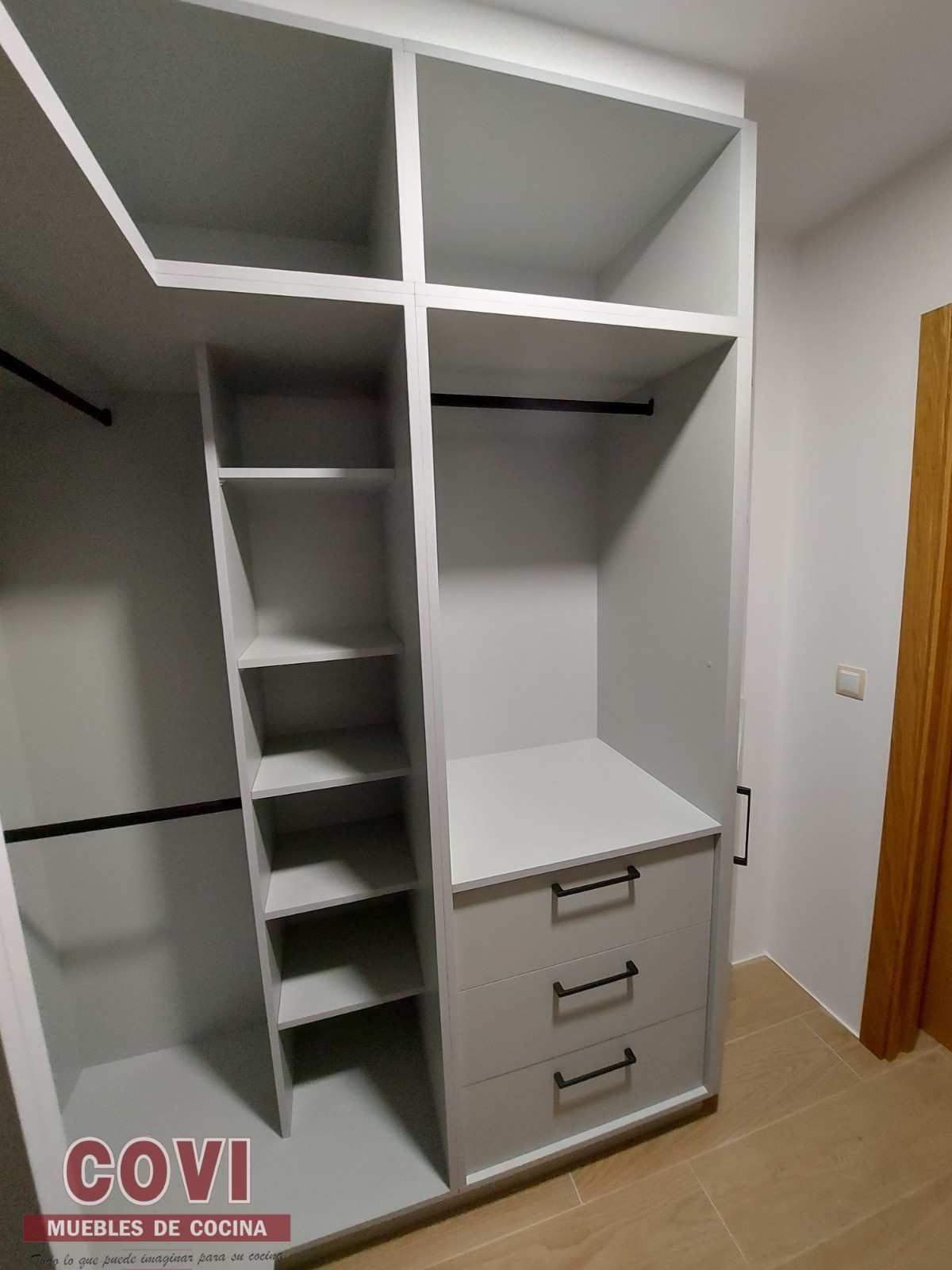 Armario empotrado gris con estantes, cajones y barras para colgar en una habitación de color claro.