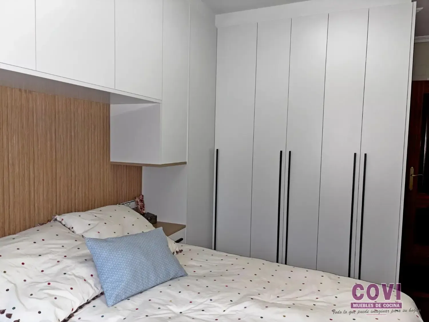 Dormitorio con cama, armario blanco y cabecero de madera clara con almacenaje incorporado.