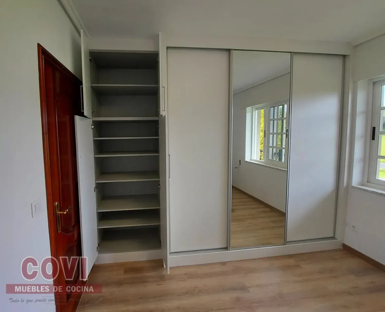 Dormitorio con armario con puertas corredizas blancas, panel de espejo y estantería alta junto a puerta marrón.