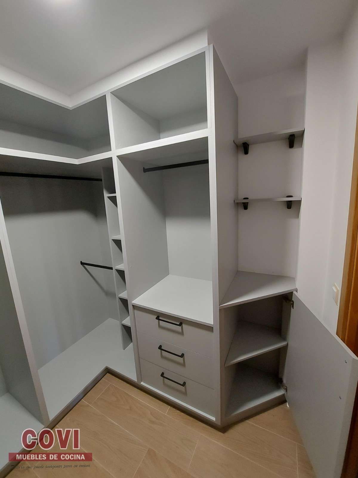 Vestidor gris con estantes, cajones y barras para colgar. Paredes y marco de puerta en tonos claros.
