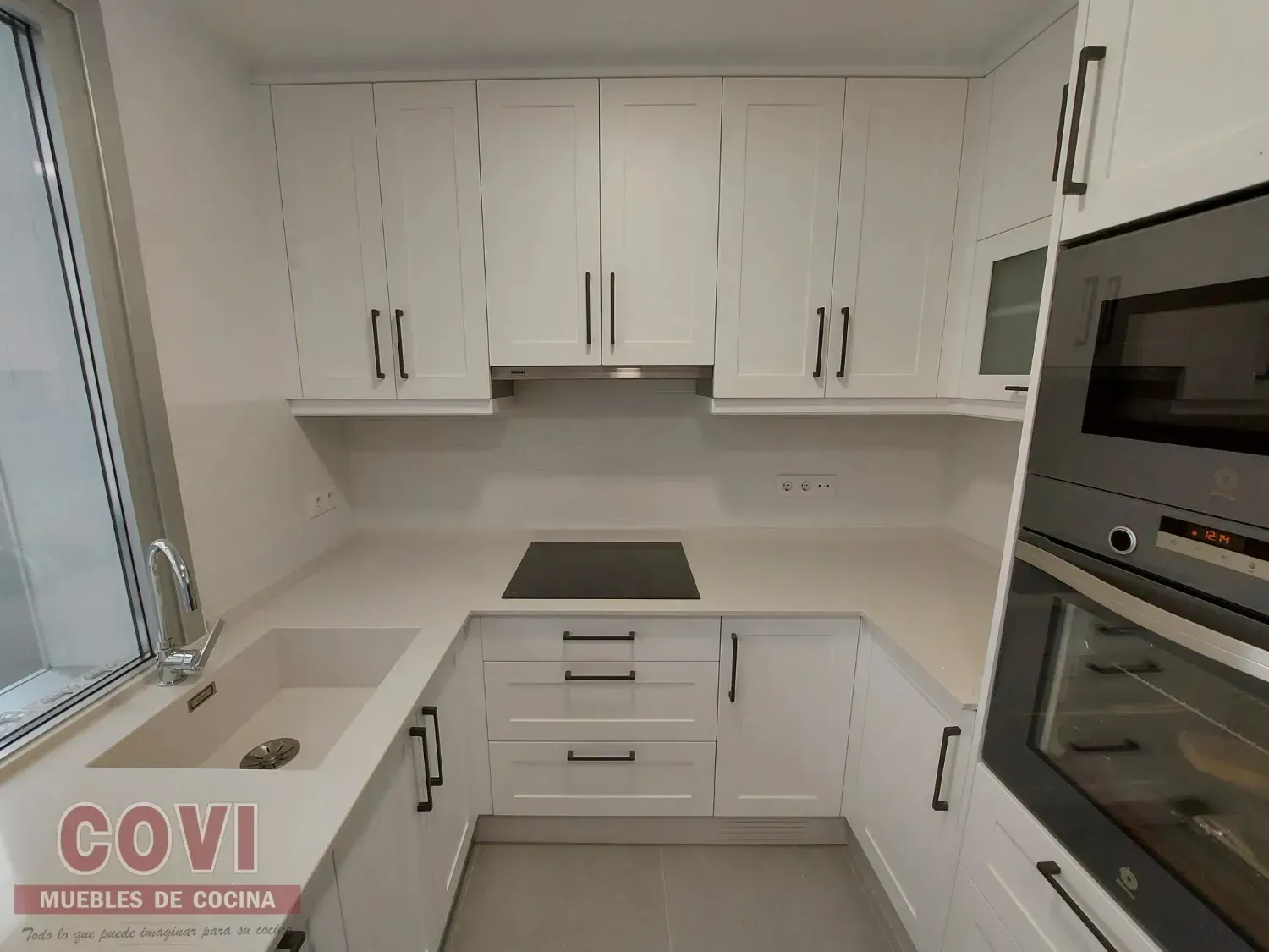 Cocina blanca con armarios altos, placa de cocina y horno empotrado. Tiradores negros y encimeras claras.