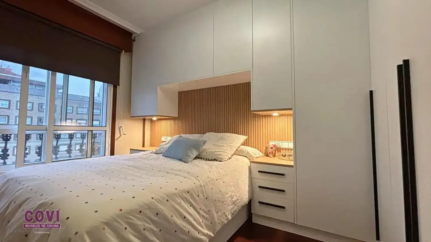 Dormitorio con armarios empotrados blancos, una cama y un gran ventanal con vistas a la ciudad.