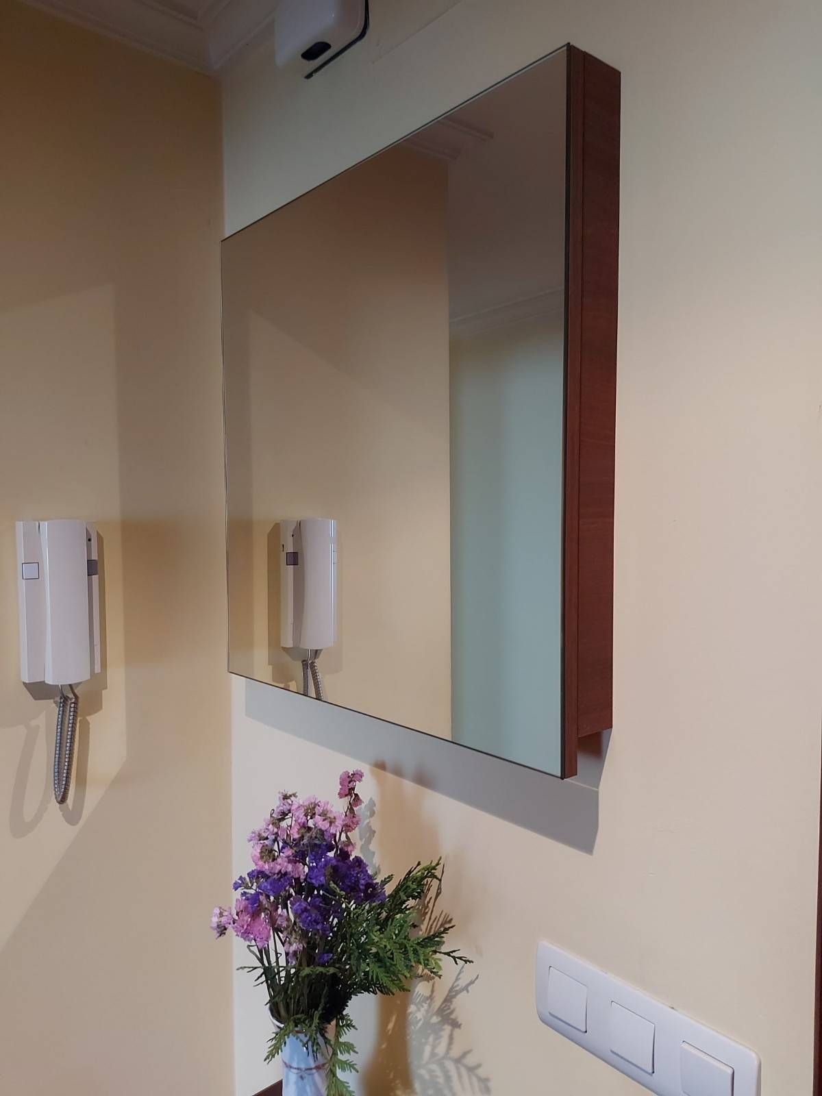 Un espejo rectangular con marco de madera marrón cuelga de una pared de color crema sobre flores en un jarrón.