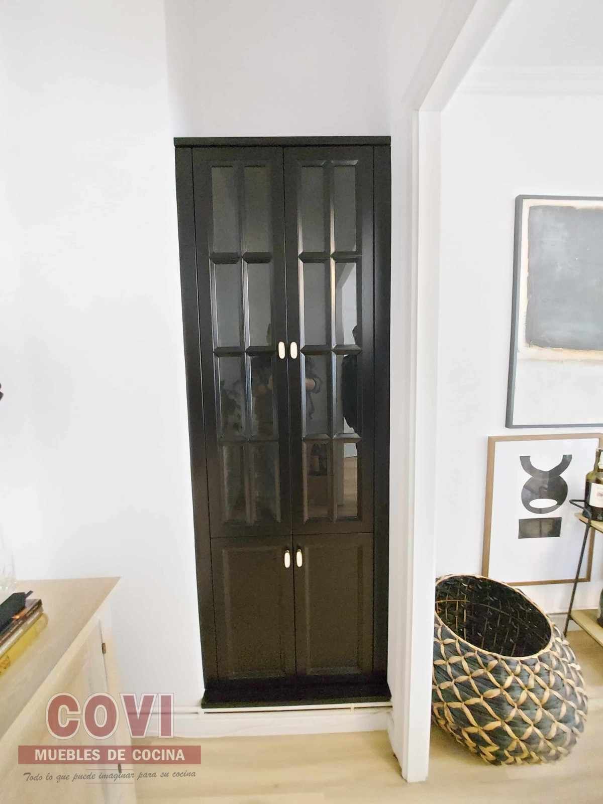 Armario negro con puertas de cristal en una habitación blanca; al lado de una puerta, una cesta y un cuadro de arte.