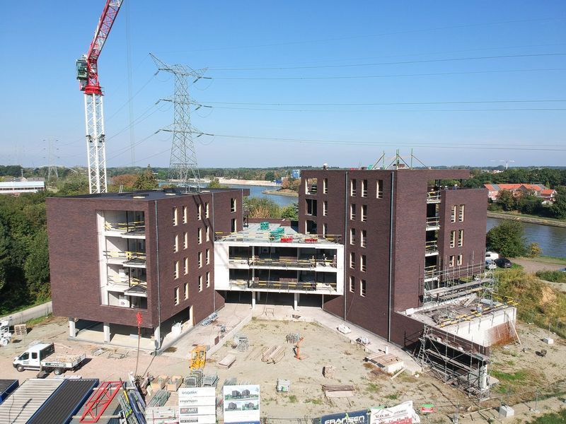 Luchtfoto van een groot gebouwencomplex in aanbouw met kranen en steigers, omgeven door een open terrein.