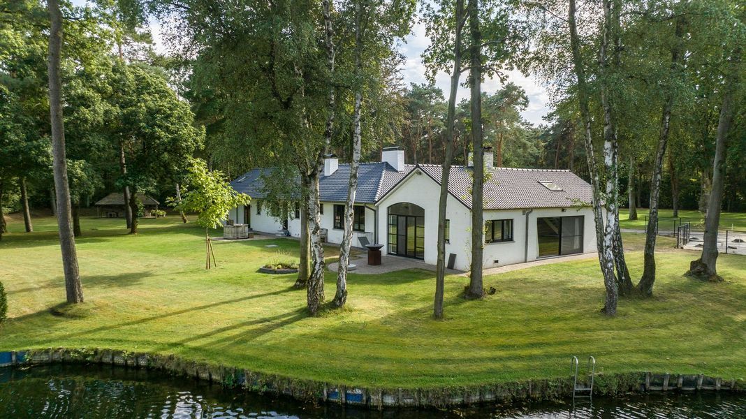 Wit huisje aan een vijver in een grasrijke, bosrijke tuin met hoge bomen.