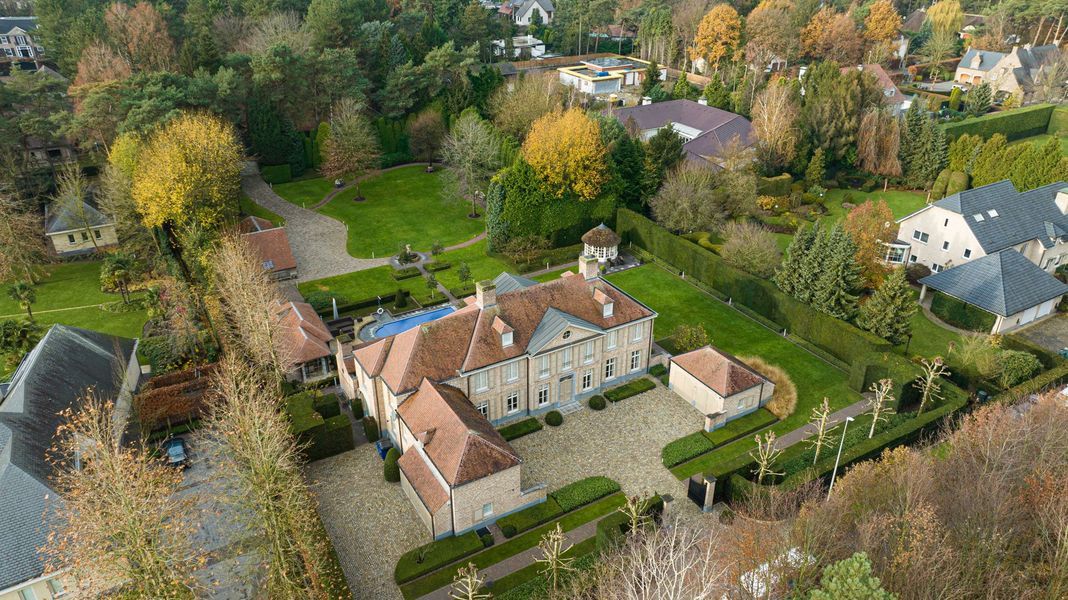 Luchtfoto van een groot wit landhuis met rode daken, omgeven door bomen en tuinen in de herfst.