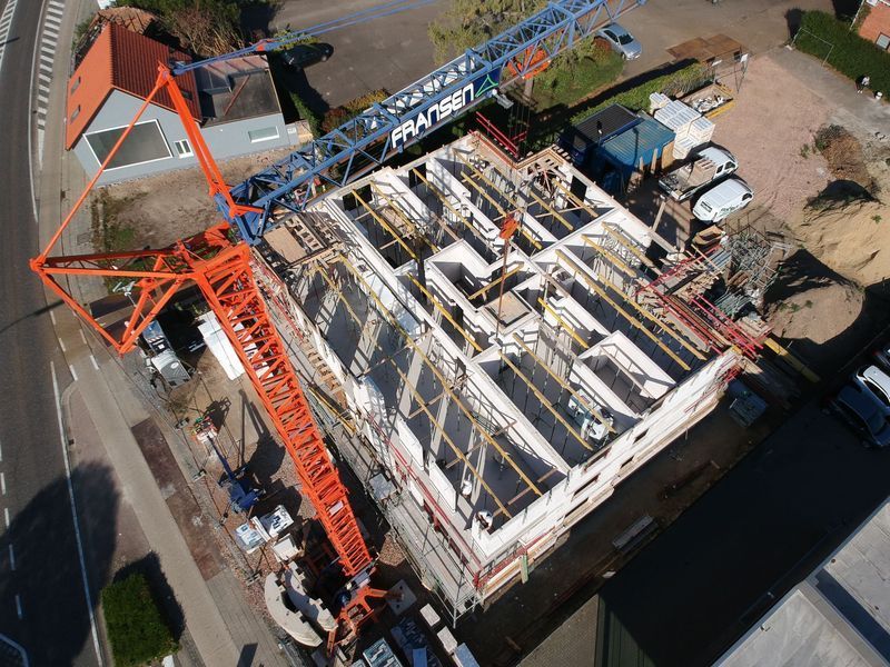 Luchtfoto van een bouwplaats met een oranje kraan die materialen over een betonnen gebouwframe hijst.