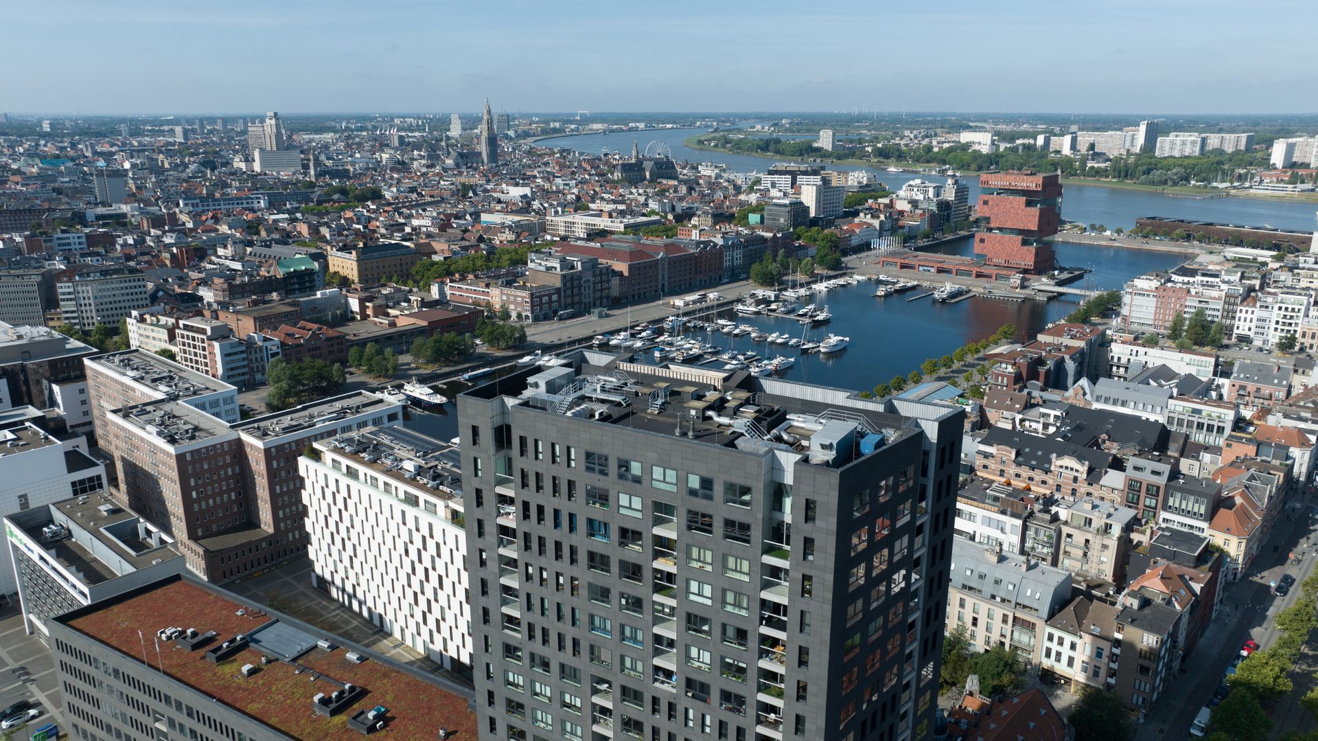 Luchtfoto van een stadsgezicht met dichtbebouwde gebouwen, een waterkant en een rode toren nabij het centrum onder een heldere hemel.
