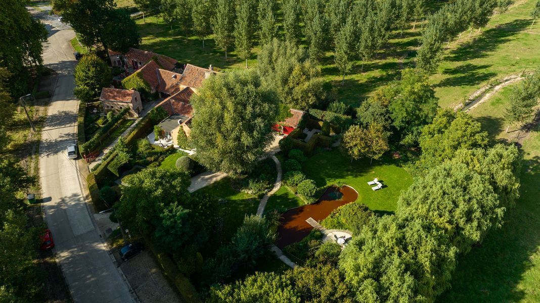 Luchtfoto van een huis met een rood dak, omgeven door weelderige bomen en een groene tuin langs een weg.