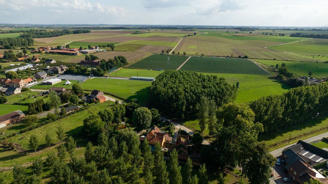 Luchtfoto van groene landbouwgrond, verspreide huizen en bomen onder een heldere hemel.