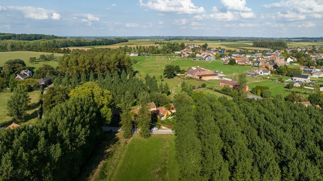 Luchtfoto van een landelijk dorp met groene velden, dichte bomen en huizen onder een gedeeltelijk bewolkte hemel.
