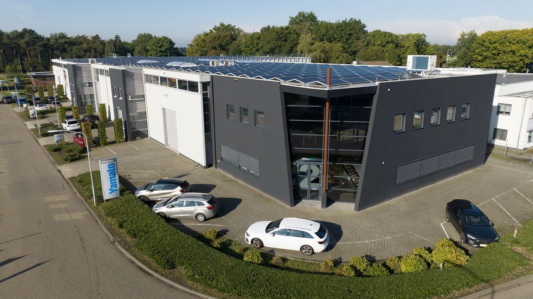 Luchtfoto van een modern, grijs industrieel gebouw met geparkeerde auto's en bomen in de buurt.