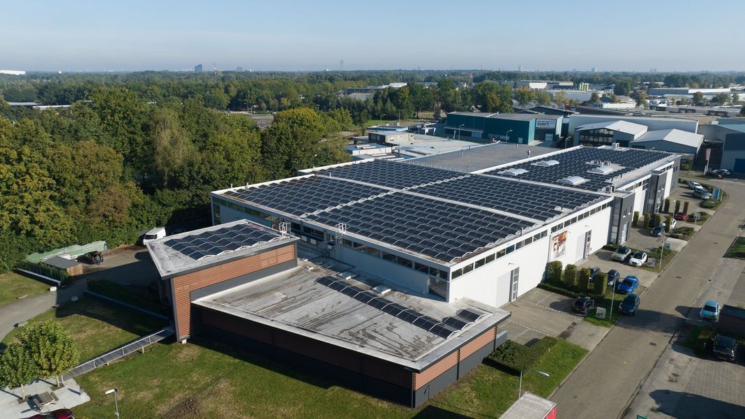 Luchtfoto van een industrieel gebouw met zonnepanelen, met bomen en een parkeerplaats in de buurt.